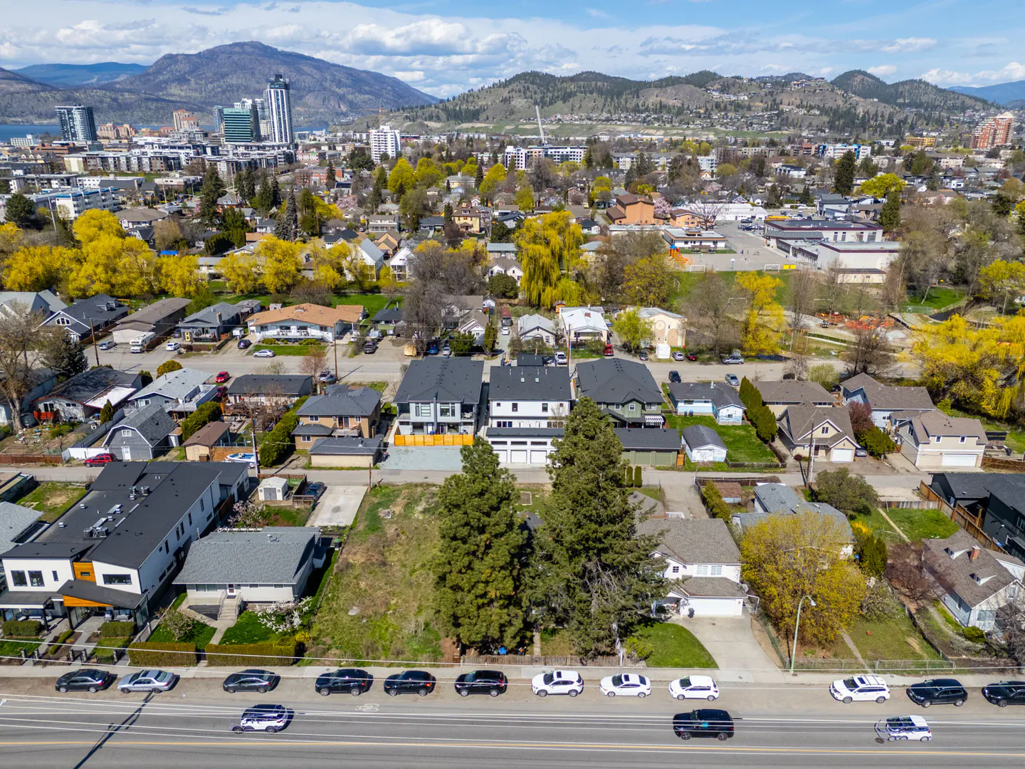 Aerial view of a residential area with houses, trees, and cars, with mountains and city buildings in the background.