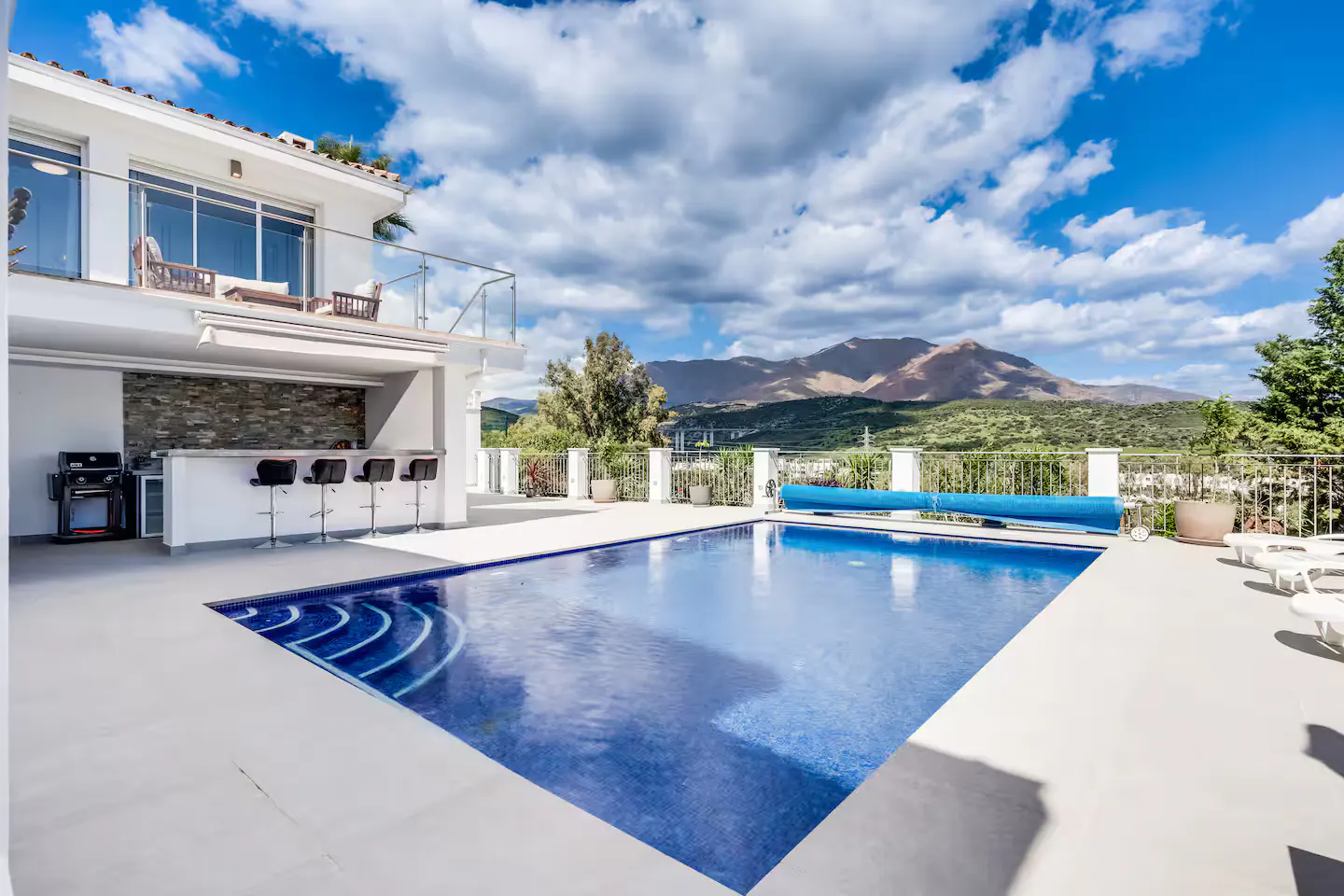 Luxury home exterior with a blue tiled pool, outdoor bar with stools, and mountain views under a cloudy sky.