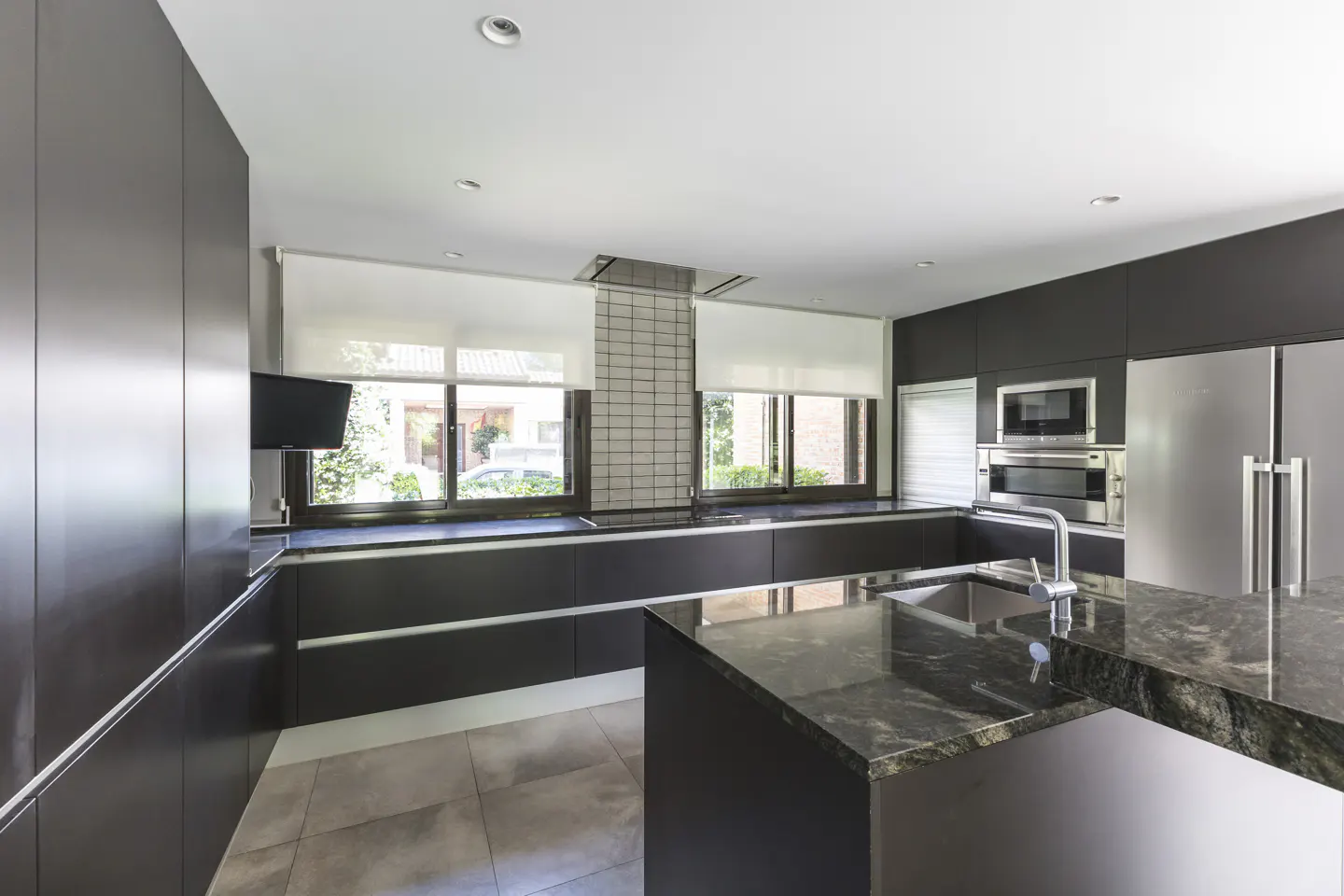 Modern kitchen with dark cabinets, stainless steel appliances, and a marble island. Windows with white shades provide natural light.