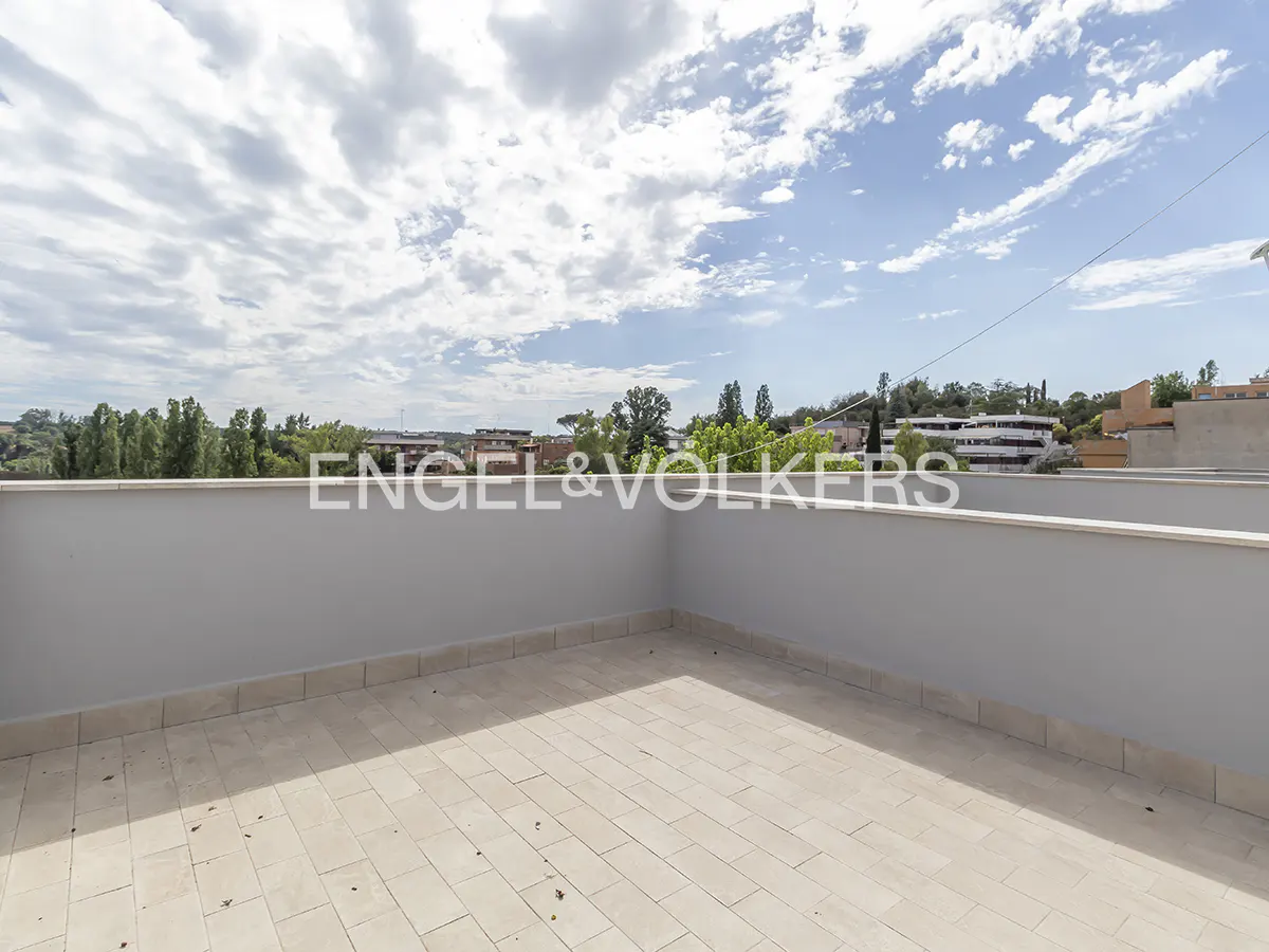 Spacious rooftop terrace with light-colored tiled flooring, grey walls, and a panoramic view of trees and distant buildings.