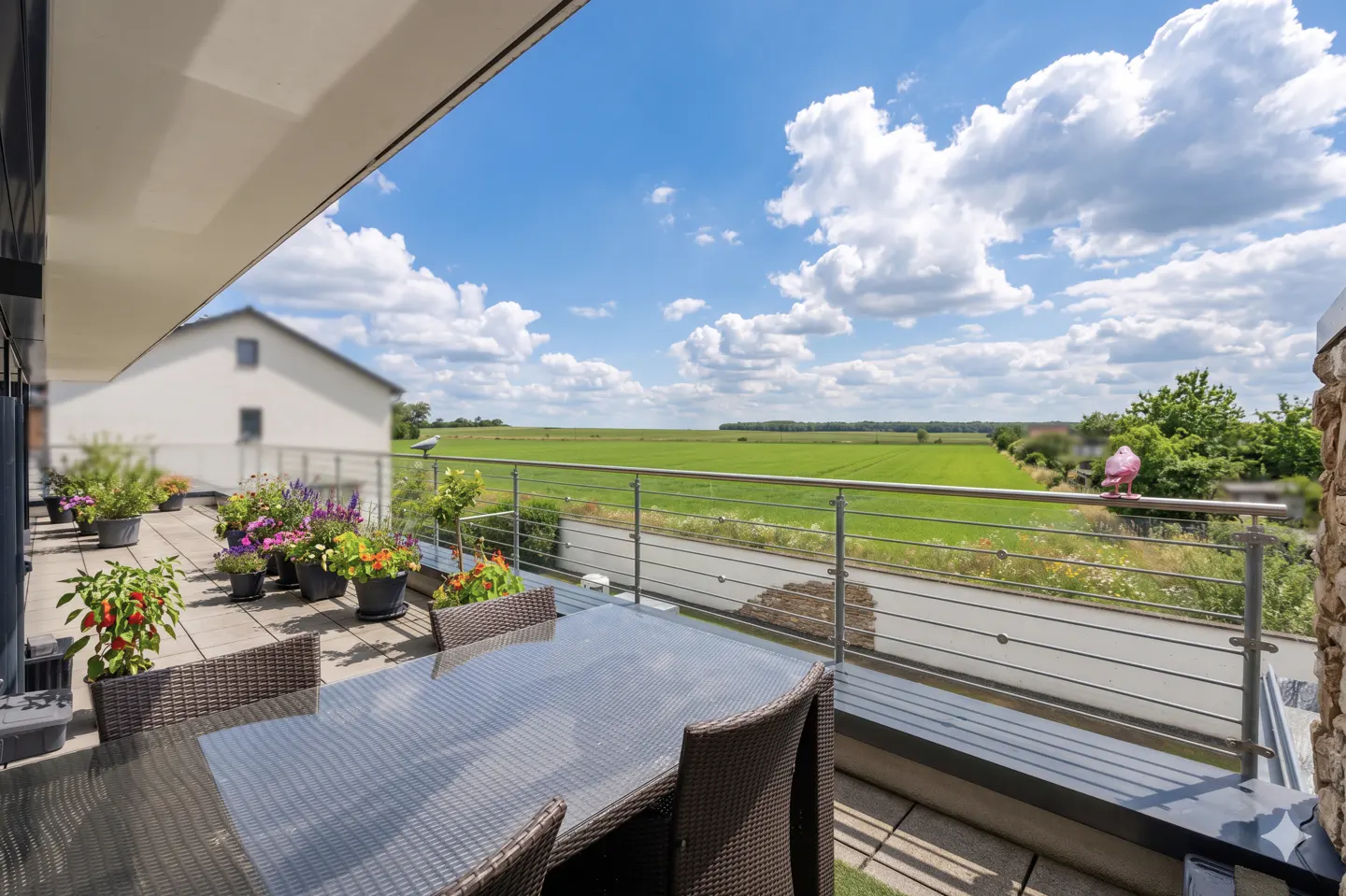 Balcony view with a table, chairs, and potted plants overlooking a green field under a blue sky with white clouds.