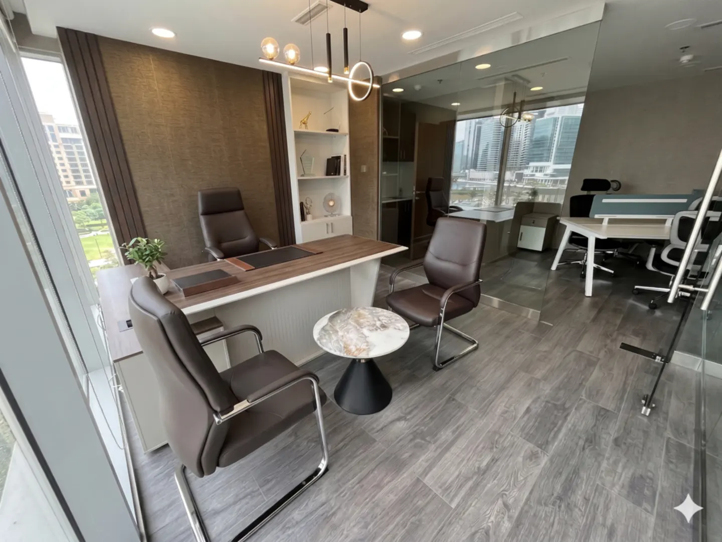 A modern office space with a large desk, brown leather chairs, and a marble-topped table. A glass wall separates the office from a larger workspace.