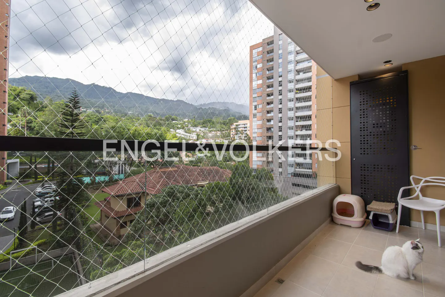 Balcony view with safety netting, showing a white cat, cat furniture, and a cityscape with mountains in the background.