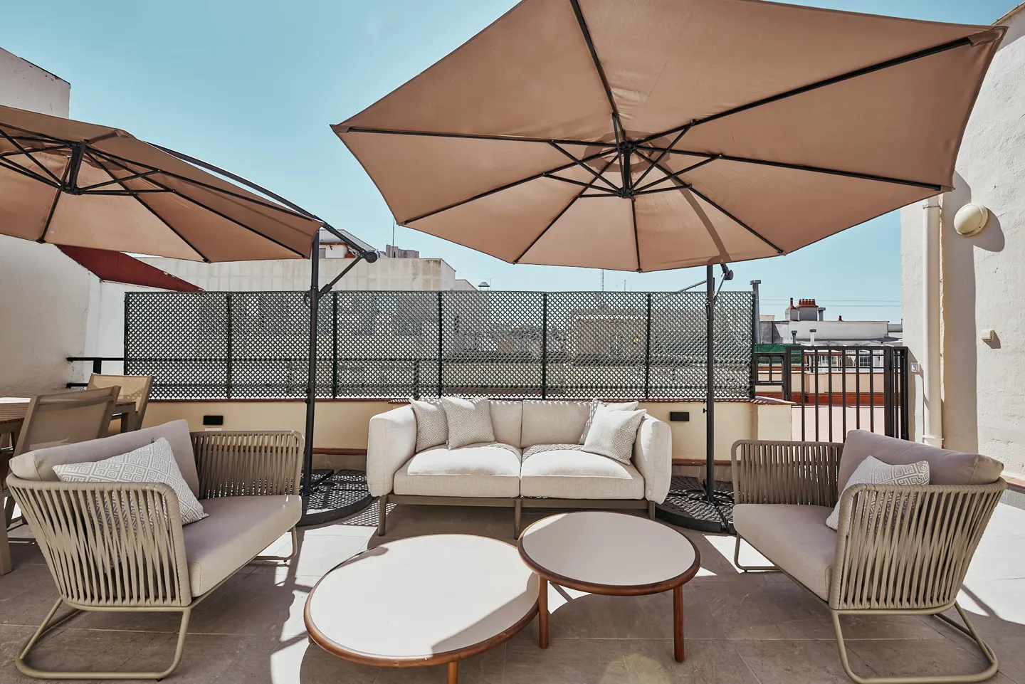 Outdoor patio with beige sofa, two chairs, and two round tables under large umbrellas, with a black metal fence in the background.