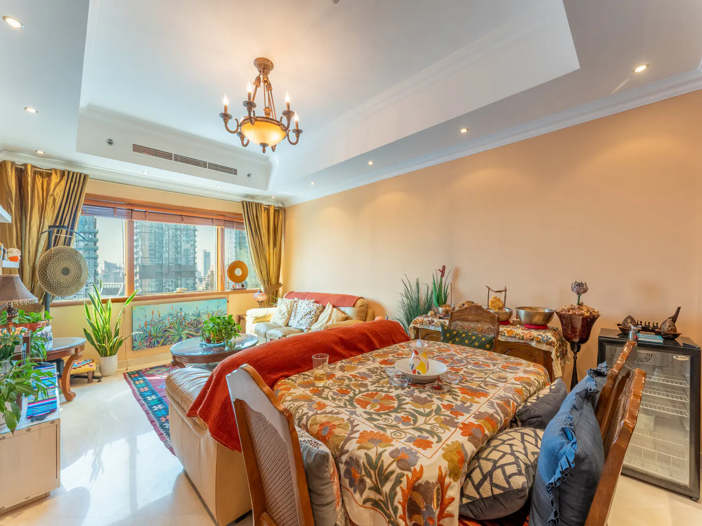 A bright living and dining area with a floral tablecloth, beige walls, and a window view of city buildings.