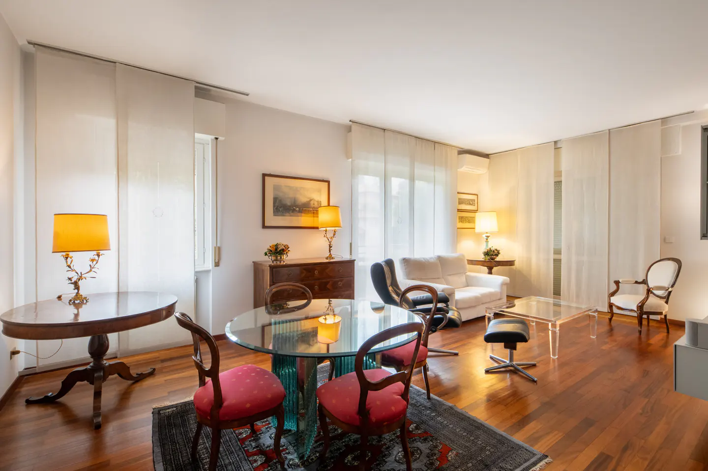 Bright living room with hardwood floors, a glass table with red chairs, and a white sofa near sheer curtains.