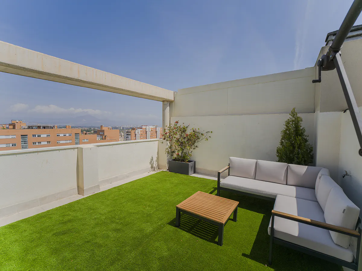 Rooftop patio with artificial grass, white sectional sofa, wood table, potted plants, and city views under a blue sky.