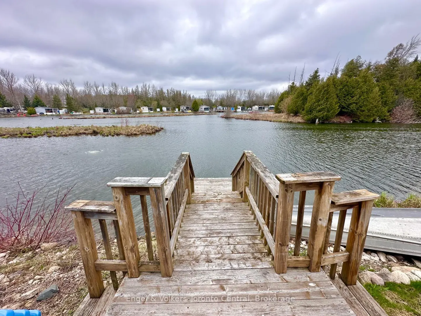 Wooden dock with railings leads to a lake under a cloudy sky. Trees and small buildings line the opposite shore.