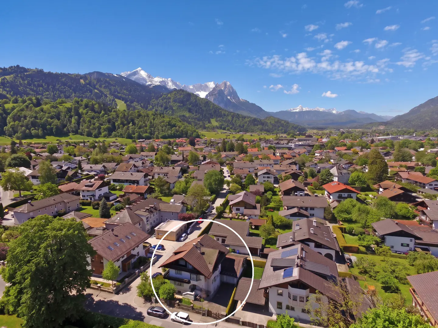 Aerial view of a house with a brown roof, surrounded by trees and other houses, with mountains in the background.