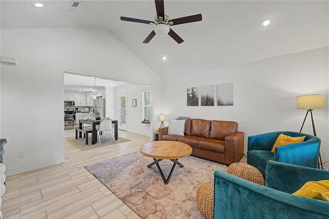 Bright living room with brown leather sofa, teal chairs, and a round wooden coffee table on a patterned rug. Open to kitchen and dining area.