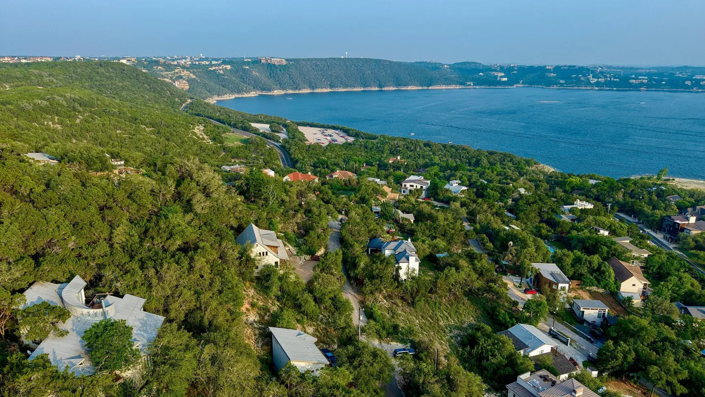 Aerial view of homes nestled in lush green trees overlooking a blue lake under a clear sky.