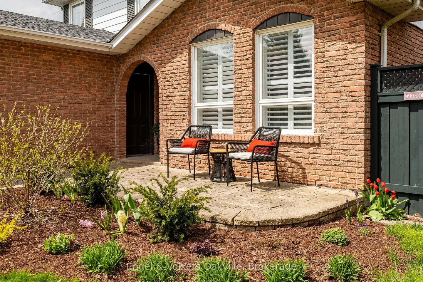 Brick house exterior with two chairs on a stone patio. A black door is framed by an archway. Landscaped garden in front.