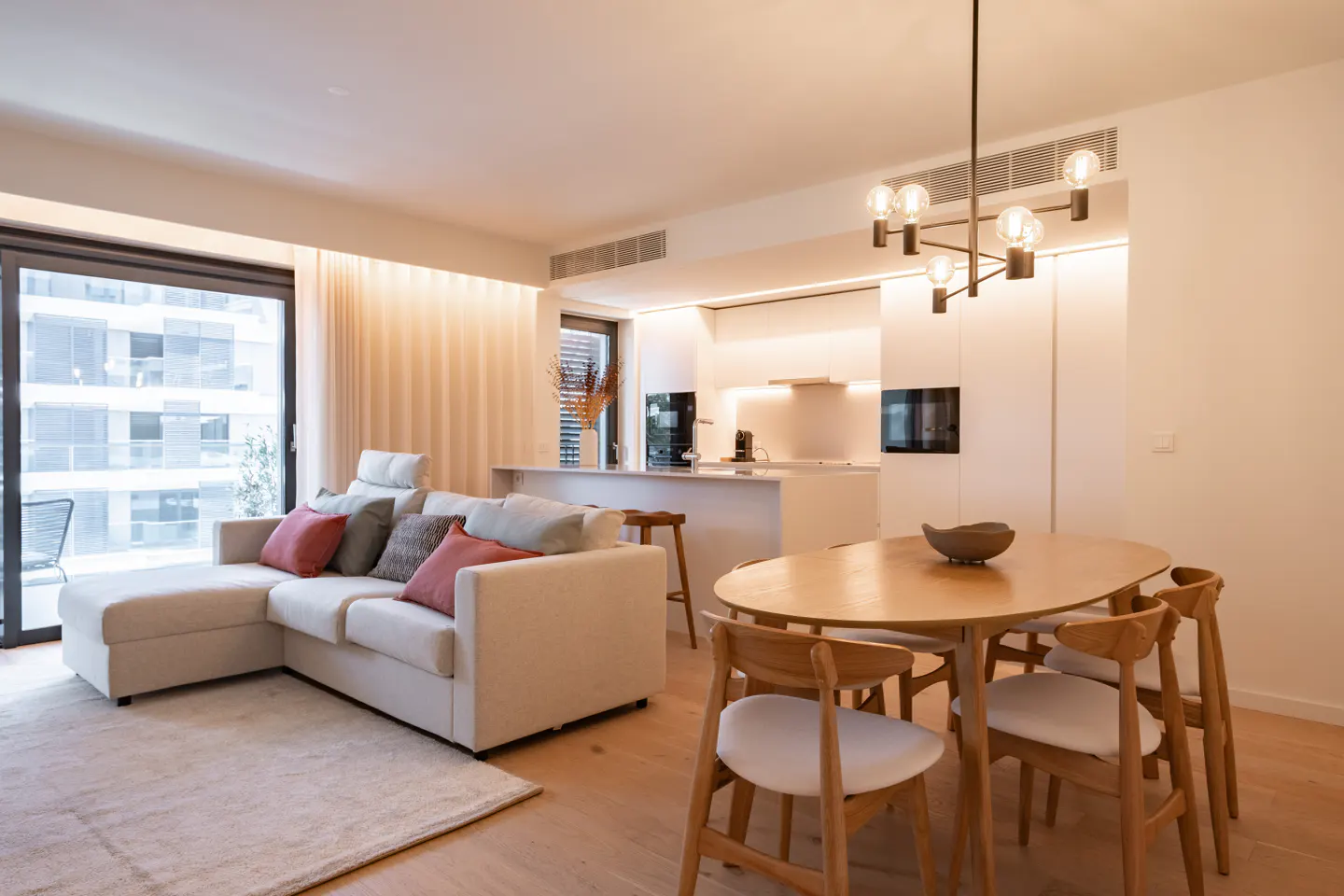 Modern open-plan living area with a white sectional sofa, wooden dining table, and minimalist kitchen in a bright space.