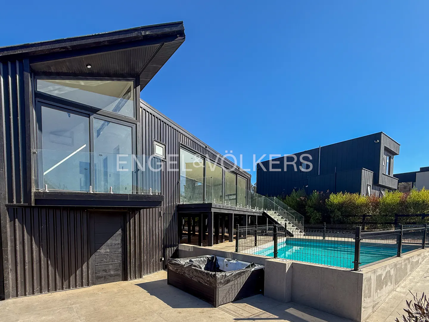 Modern black house with a pool and hot tub under a clear blue sky. Engel & Völkers logo on the house.