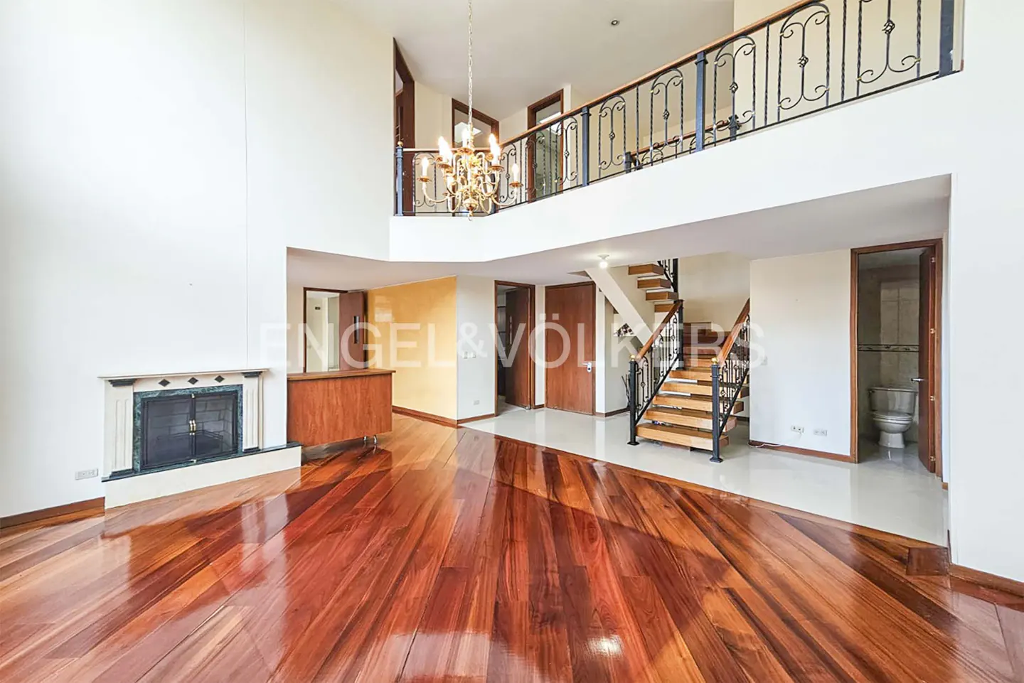 A bright, open-concept living room with hardwood floors, a fireplace, and a staircase leading to a second-floor balcony.
