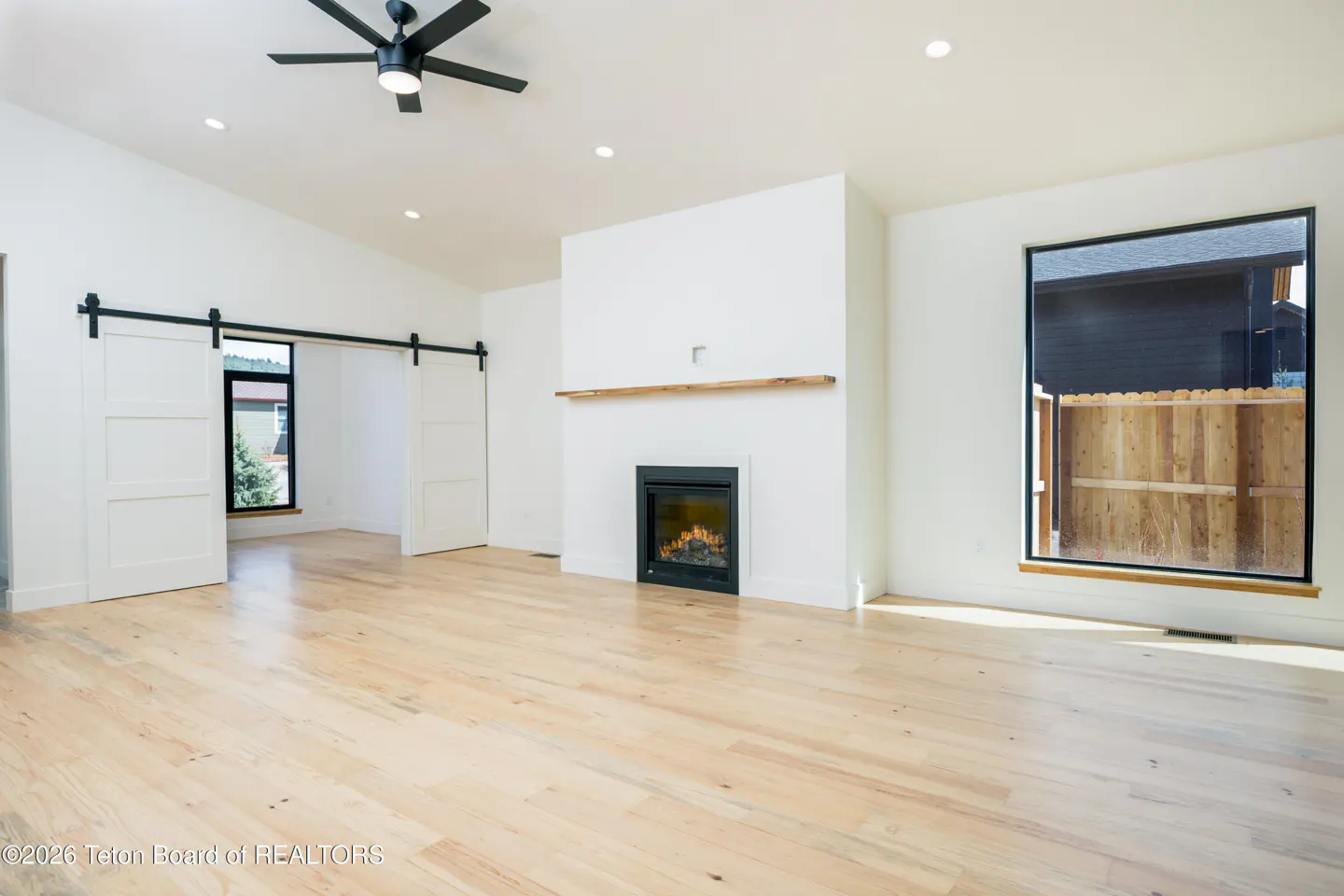 Bright, empty living room with light wood floors, white walls, and a black ceiling fan. A fireplace and sliding barn doors add character.
