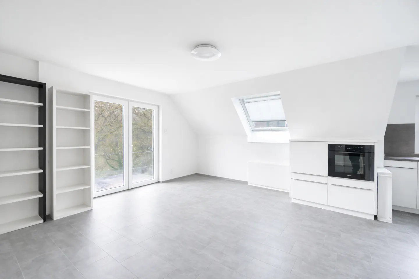 Bright, empty attic apartment with gray tile floor, white walls, and built-in white kitchen cabinets with black oven. Bookshelves and glass doors to balcony.