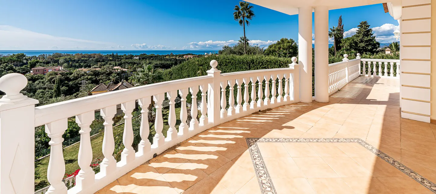 View from a sunny balcony with white balustrade overlooking lush greenery and the ocean under a blue sky.