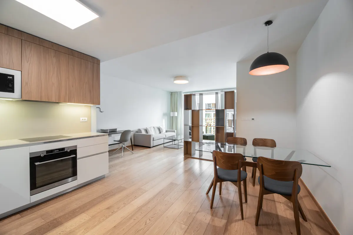 Open-concept apartment with wood floors, white walls, and modern furniture. Kitchen, dining, and living areas are visible.