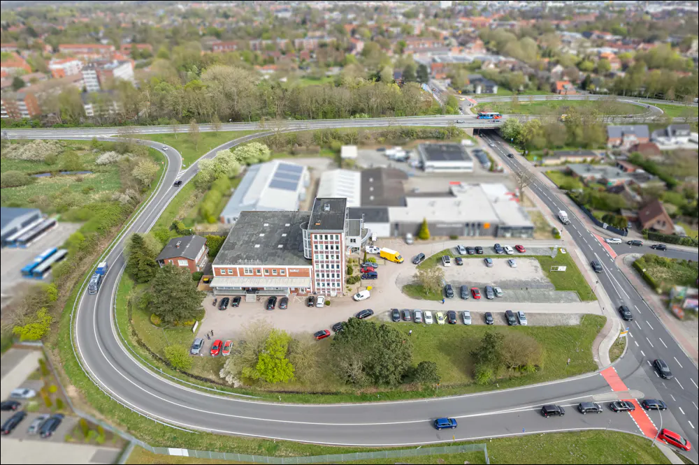 Aerial view of a brick building with a tower, surrounded by parking, roads, and greenery in a suburban area.
