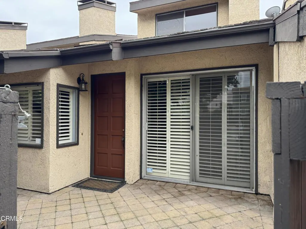 Exterior view of a tan stucco house with a brown front door, sliding glass door with white shutters, and a brick patio.