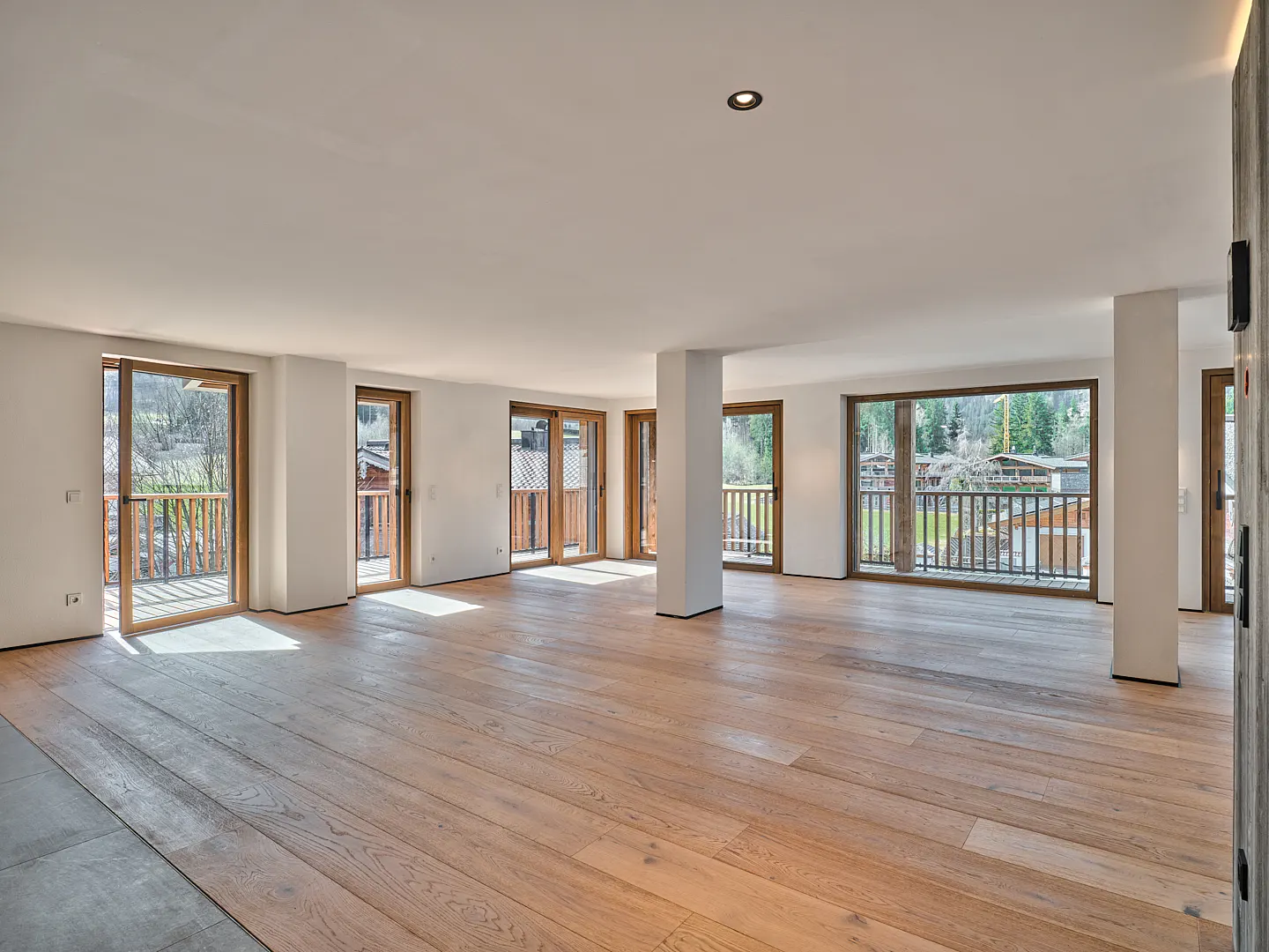 Bright, empty room with light wood floors, white walls, and multiple glass doors leading to balconies with wooden railings.