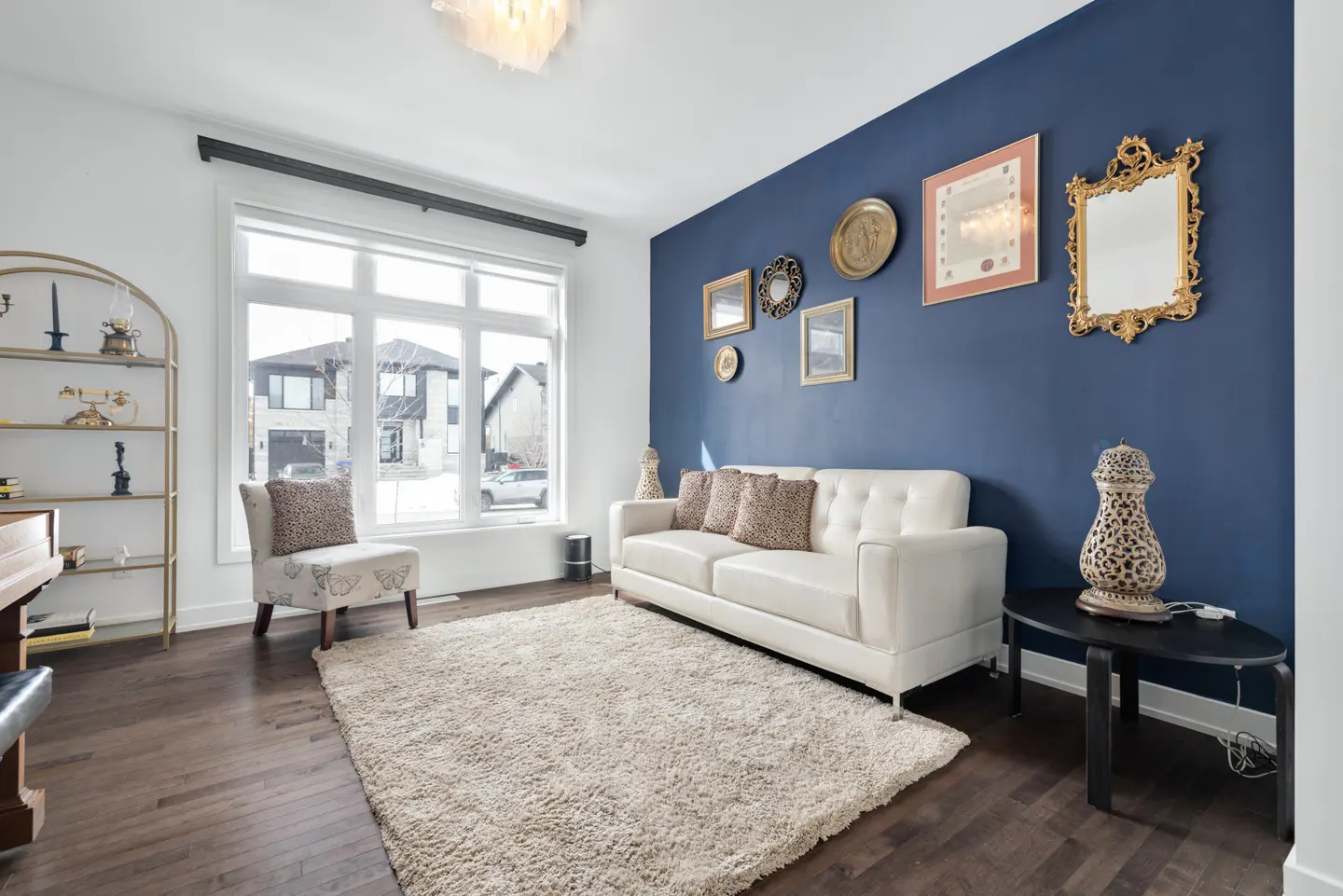 Living room with white sofa, beige rug, and blue accent wall decorated with gold-framed art. Large window with view of a house.