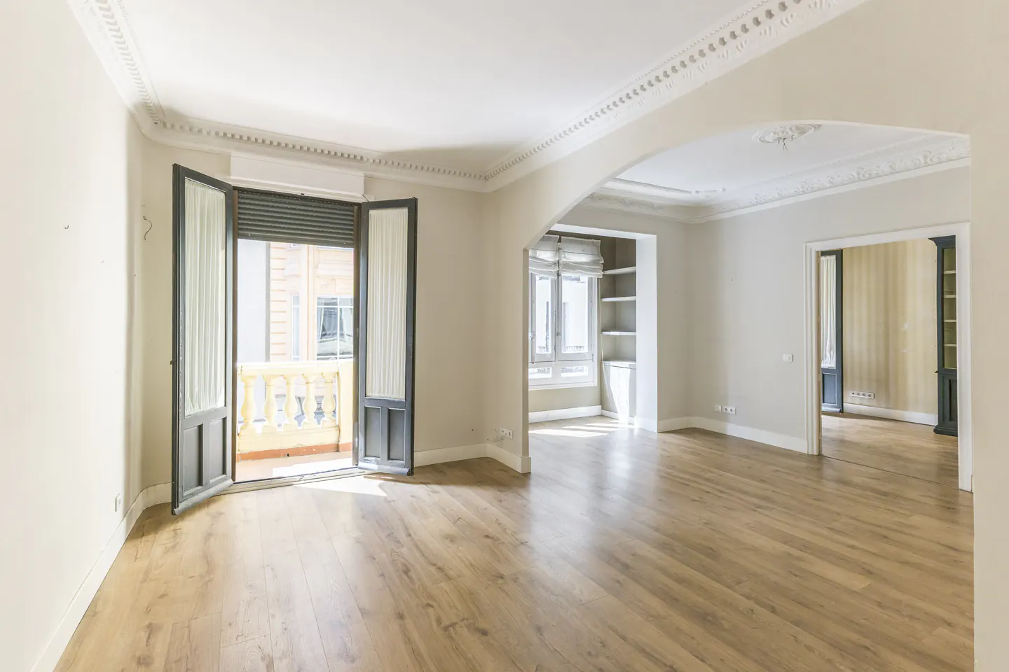 Bright, empty room with wood floors, white walls, and decorative molding. Open doors lead to a balcony with a yellow railing. Arched doorway to another room.