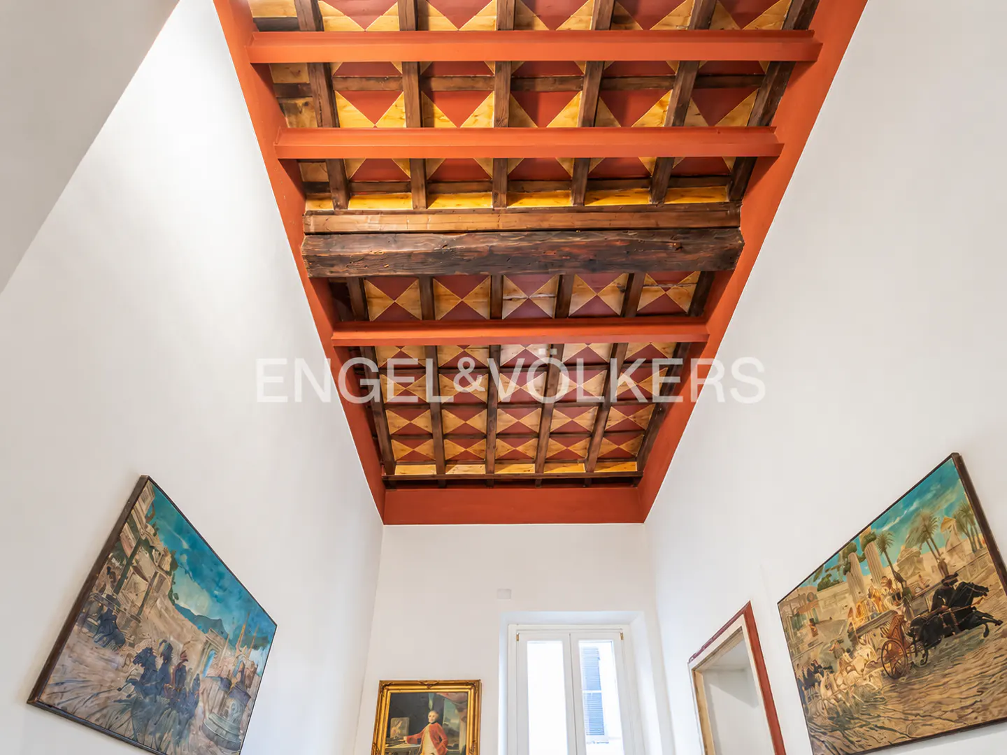Hallway with white walls, paintings, and a decorative ceiling with red and yellow geometric patterns.