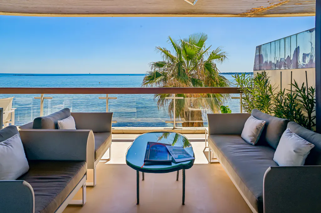 Outdoor patio with gray sofas, white pillows, and a glass table with brochures overlooking the ocean and a palm tree on a sunny day.