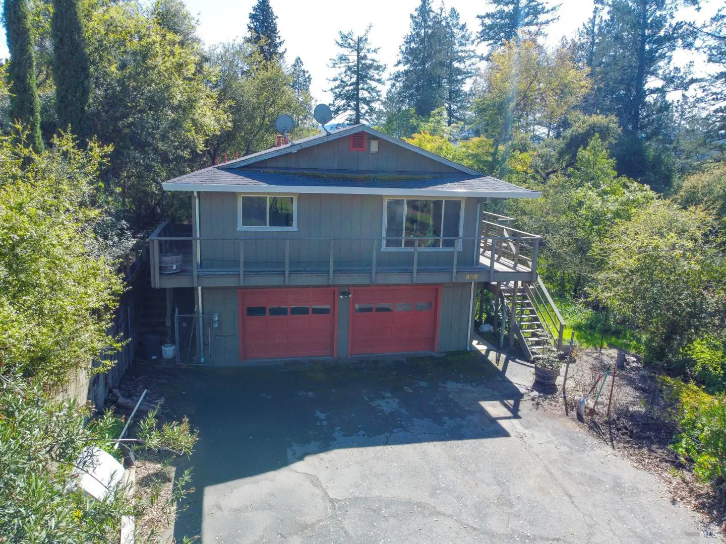 Two-story gray house with red garage doors, surrounded by trees. A wooden staircase leads to a second-floor deck.