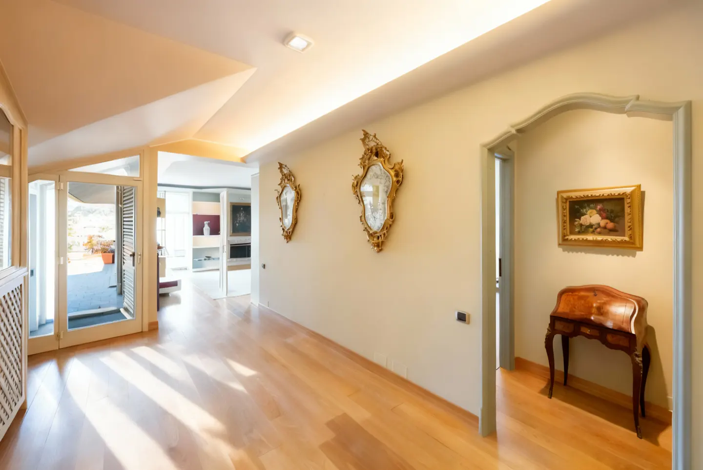 Hallway with light wood floors, cream walls, and ornate gold mirrors. A desk sits near an arched doorway with a floral painting. Glass doors lead to a balcony.