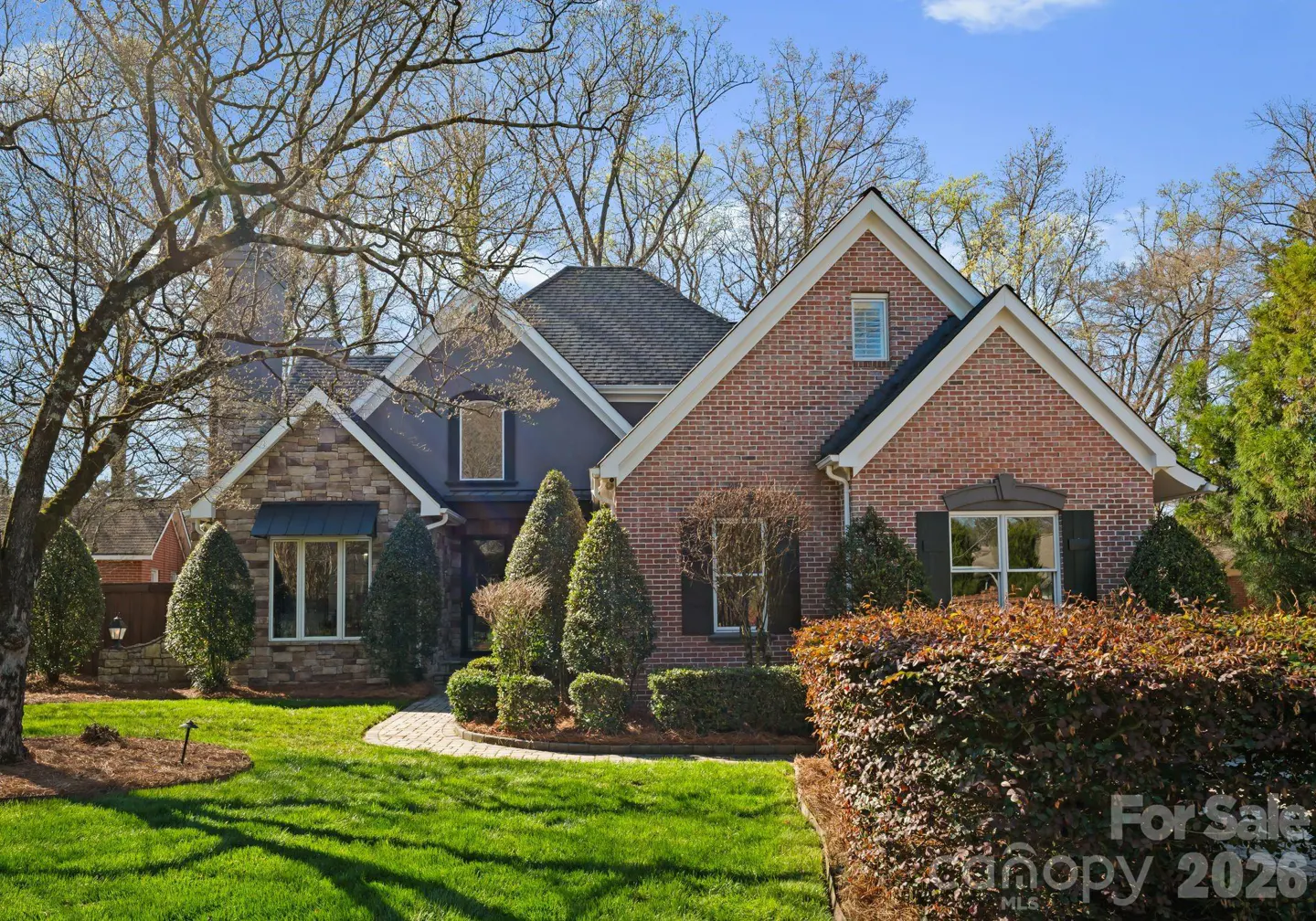 Two-story house with brick and stone facade, dark roof, green lawn, and manicured bushes under a blue sky.