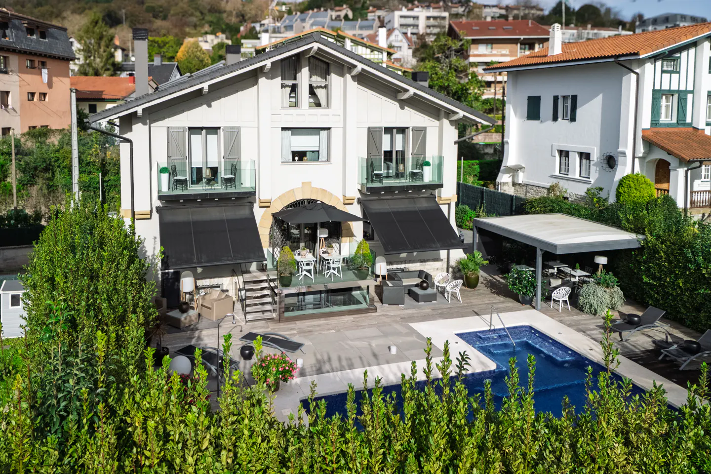 Two-story white house with a pool and patio. Black awnings cover the patio area. Green bushes in the foreground.