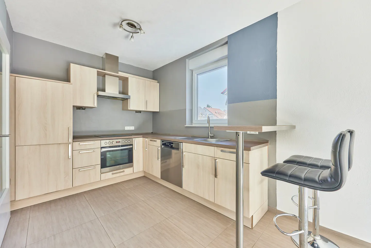A modern kitchen with light wood cabinets, stainless steel appliances, and a gray accent wall. A breakfast bar with two black stools is on the right.