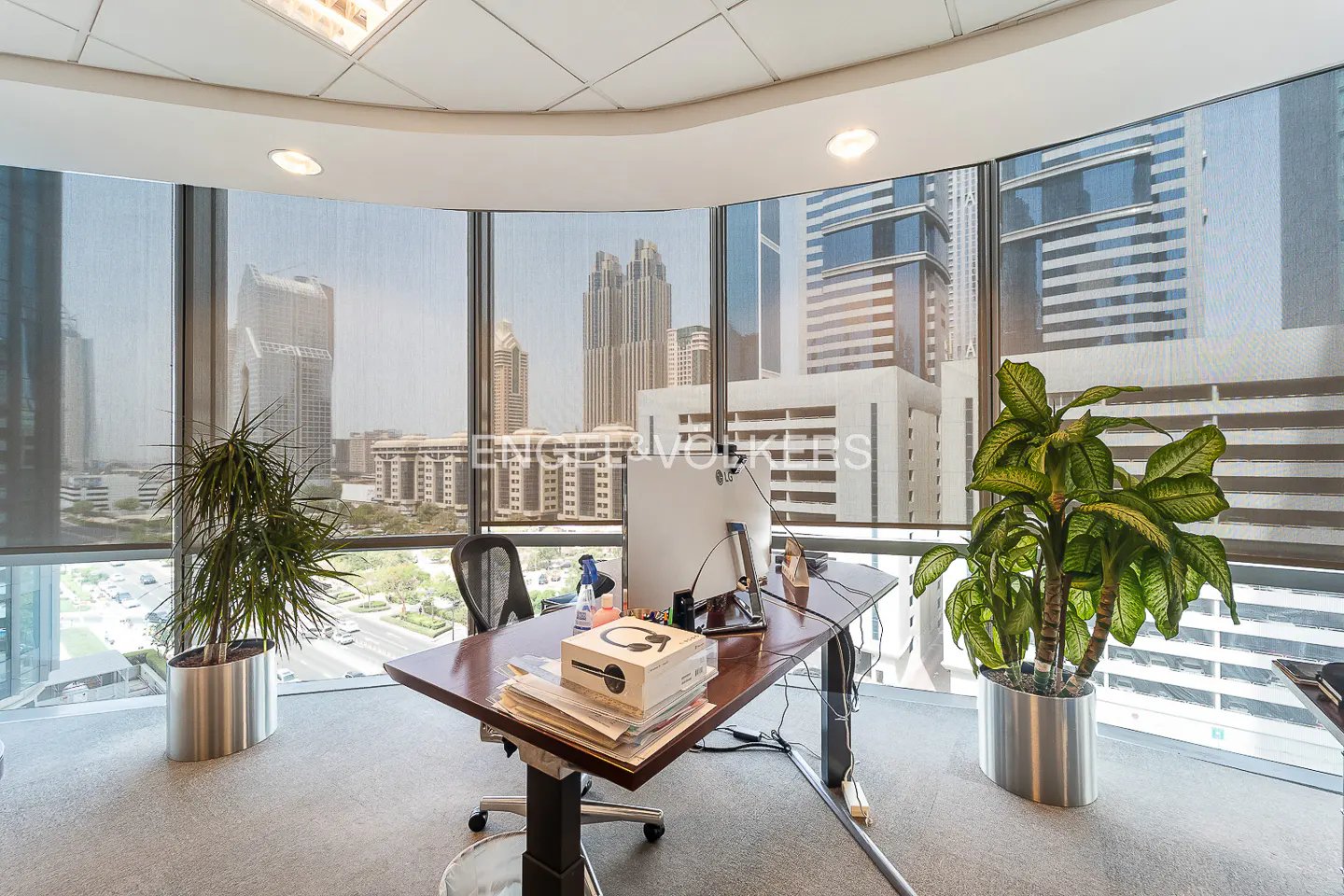 Modern office with a wooden desk, computer, and potted plants, set against a backdrop of city skyscrapers and glass windows.