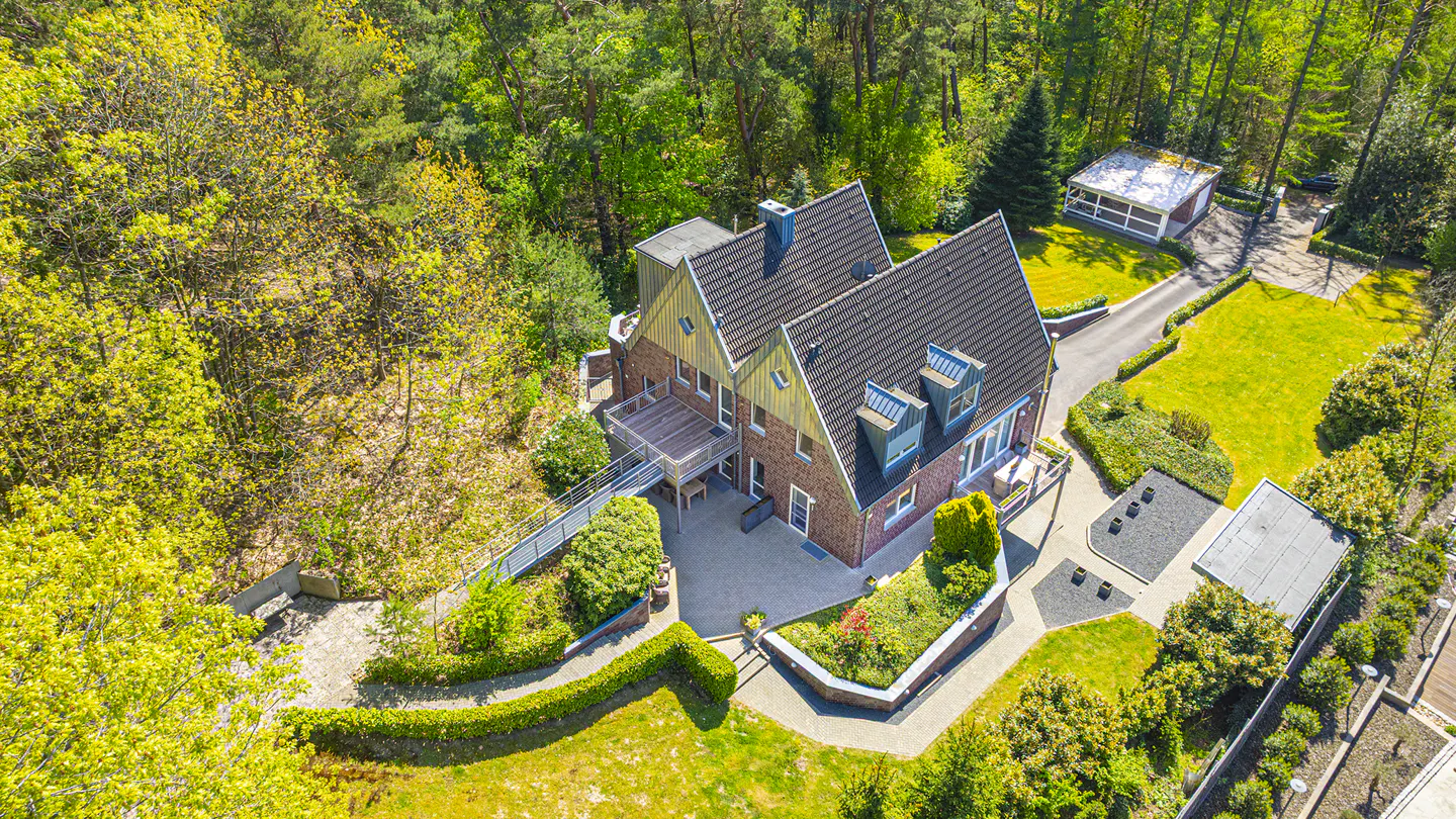 Aerial view of a brick house with a dark roof, surrounded by green trees and manicured lawns. A walkway leads to the house.