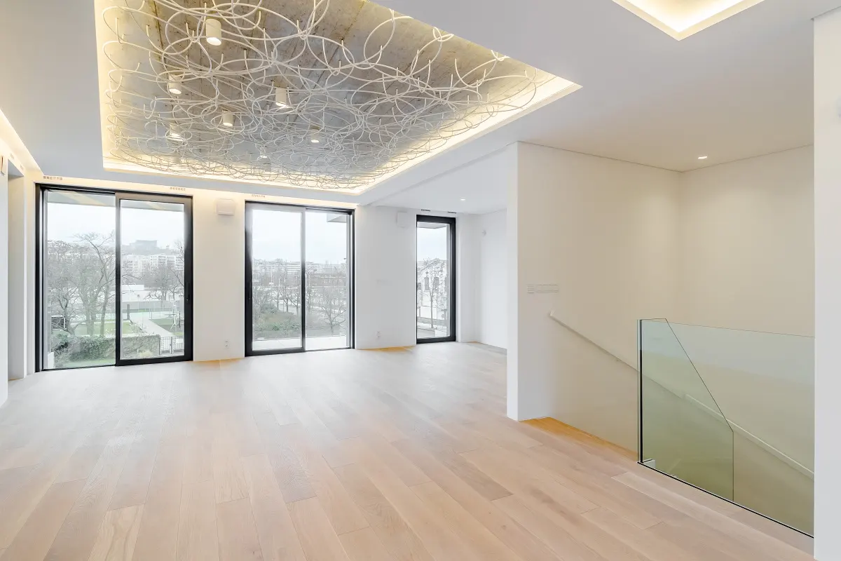 Bright, modern room with light wood floors, white walls, and large windows. A unique, artistic light fixture adorns the ceiling. Glass railing on stairs.