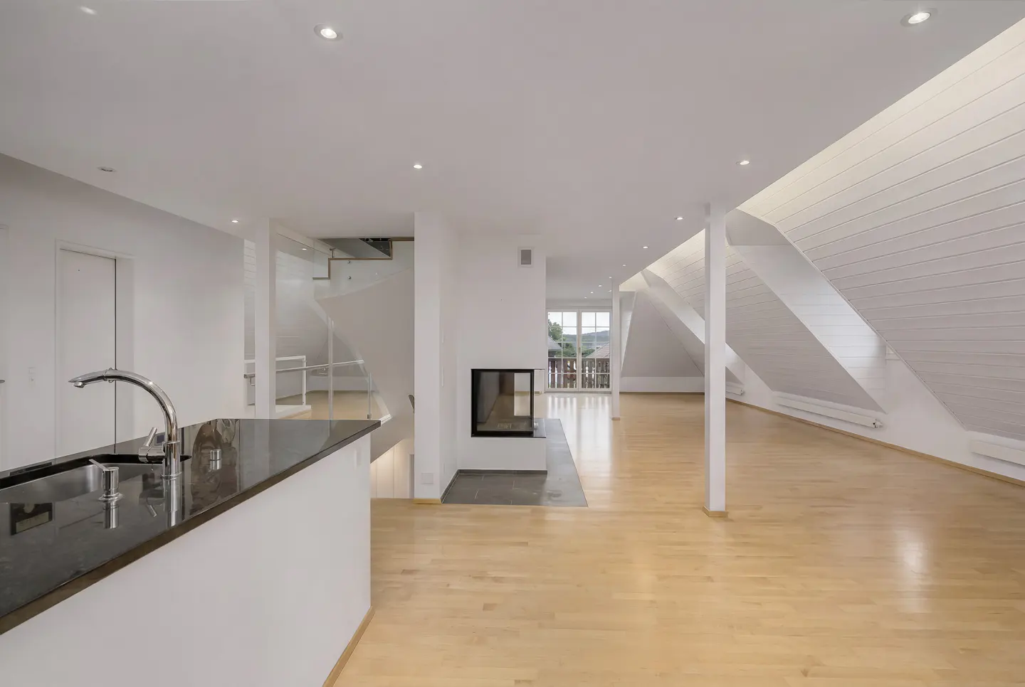 Bright, open-concept living space with wood floors, white walls, and a modern fireplace. A kitchen island with a black countertop is visible.