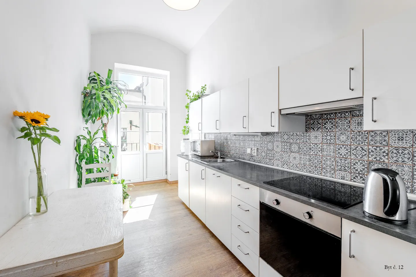 Modern white kitchen with patterned tile backsplash, wooden floors, a dining table with sunflowers, and a glass door.