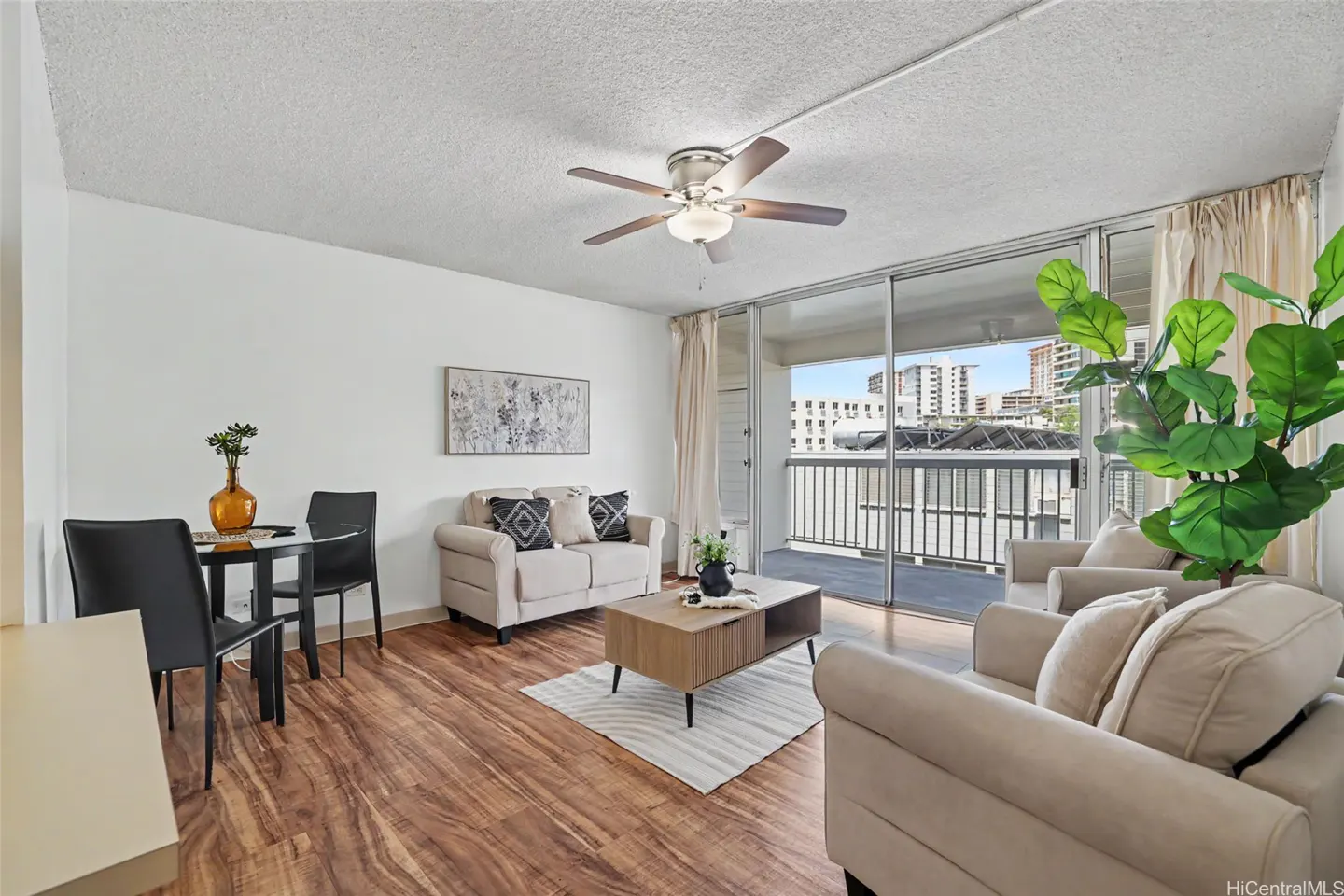 A bright living room with wood floors, white walls, and beige furniture. A ceiling fan hangs above, and a balcony offers a city view.