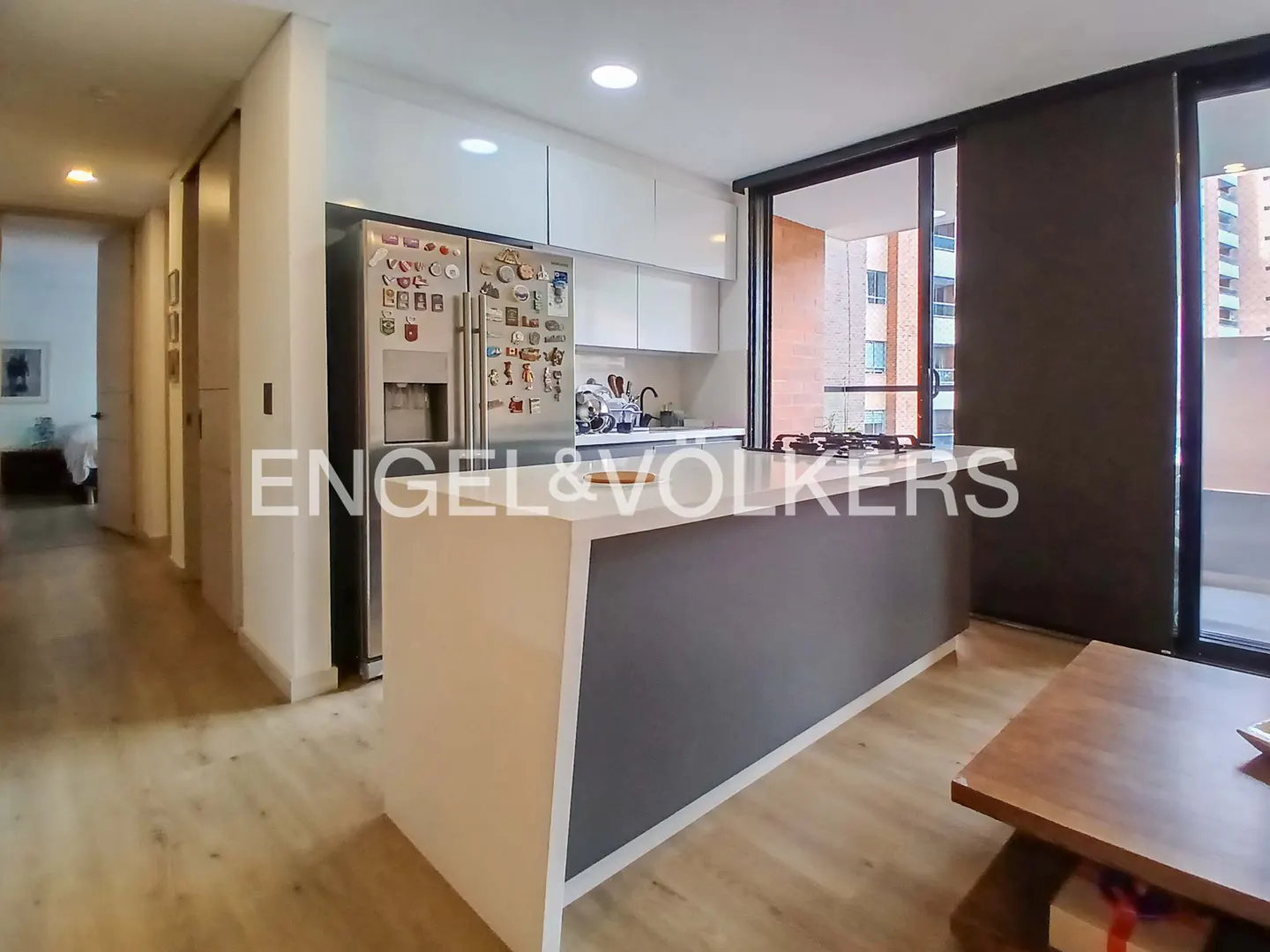 A modern kitchen with white cabinets, a stainless steel refrigerator covered in magnets, and a gray and white island. Sliding glass doors lead to a balcony.