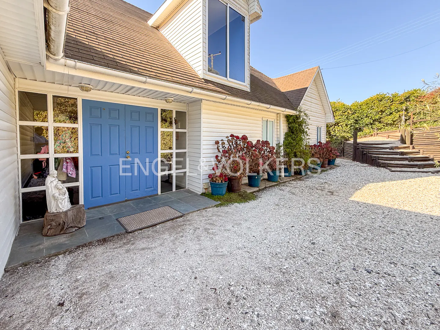 Exterior of a white house with a blue front door, gravel driveway, and potted plants.