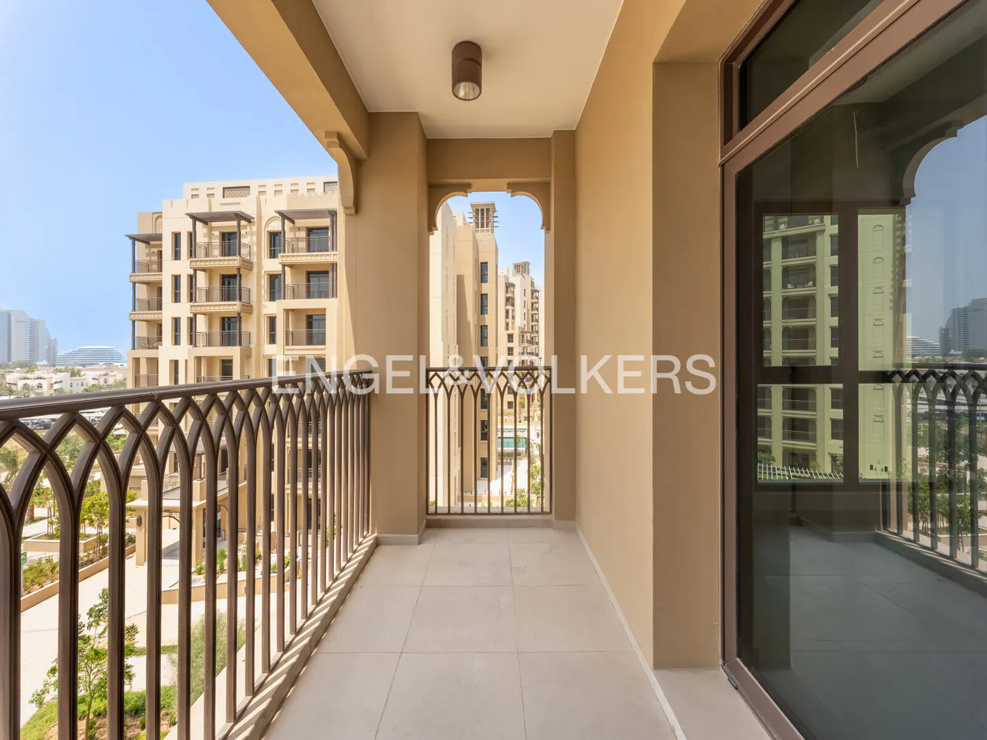 Balcony view with brown metal railings, beige tile floor, and tan building exterior. Tall buildings are visible in the background.