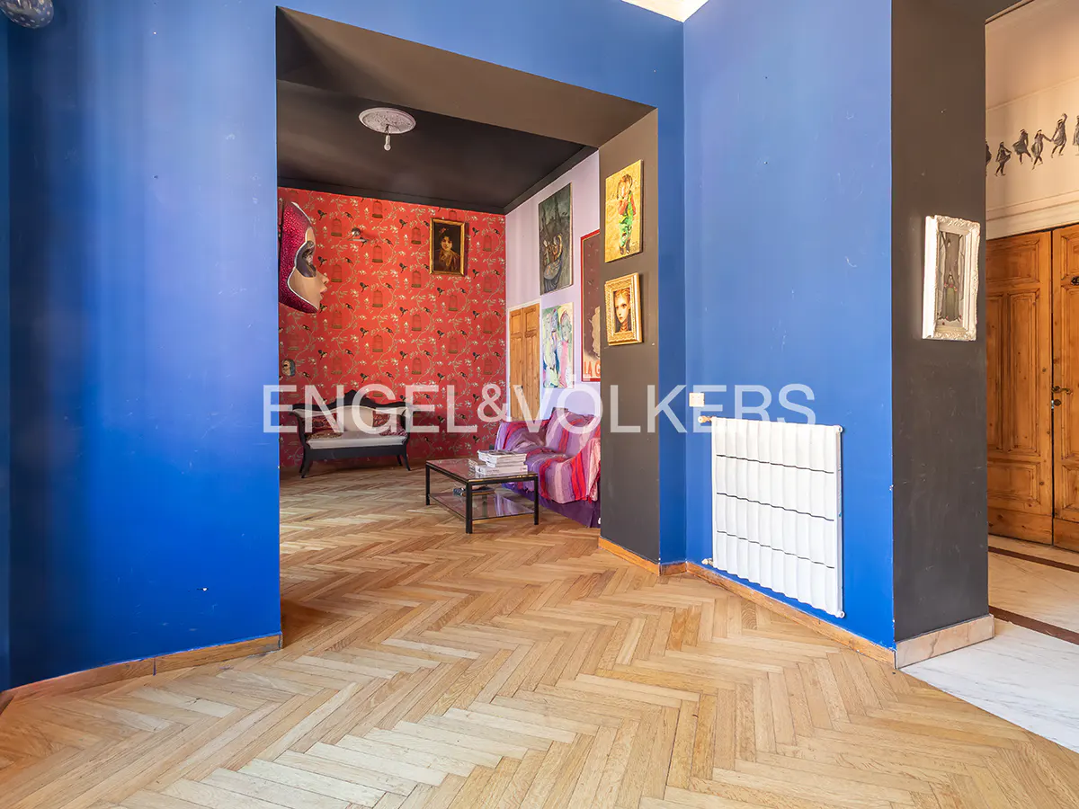 Interior view of a home with blue walls, herringbone wood floors, and a glimpse into a red-walled room with art.