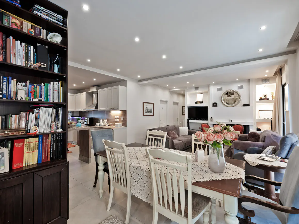 Open-concept living space with a dining table, white chairs, and a vase of pink roses. A bookcase and kitchen are visible.