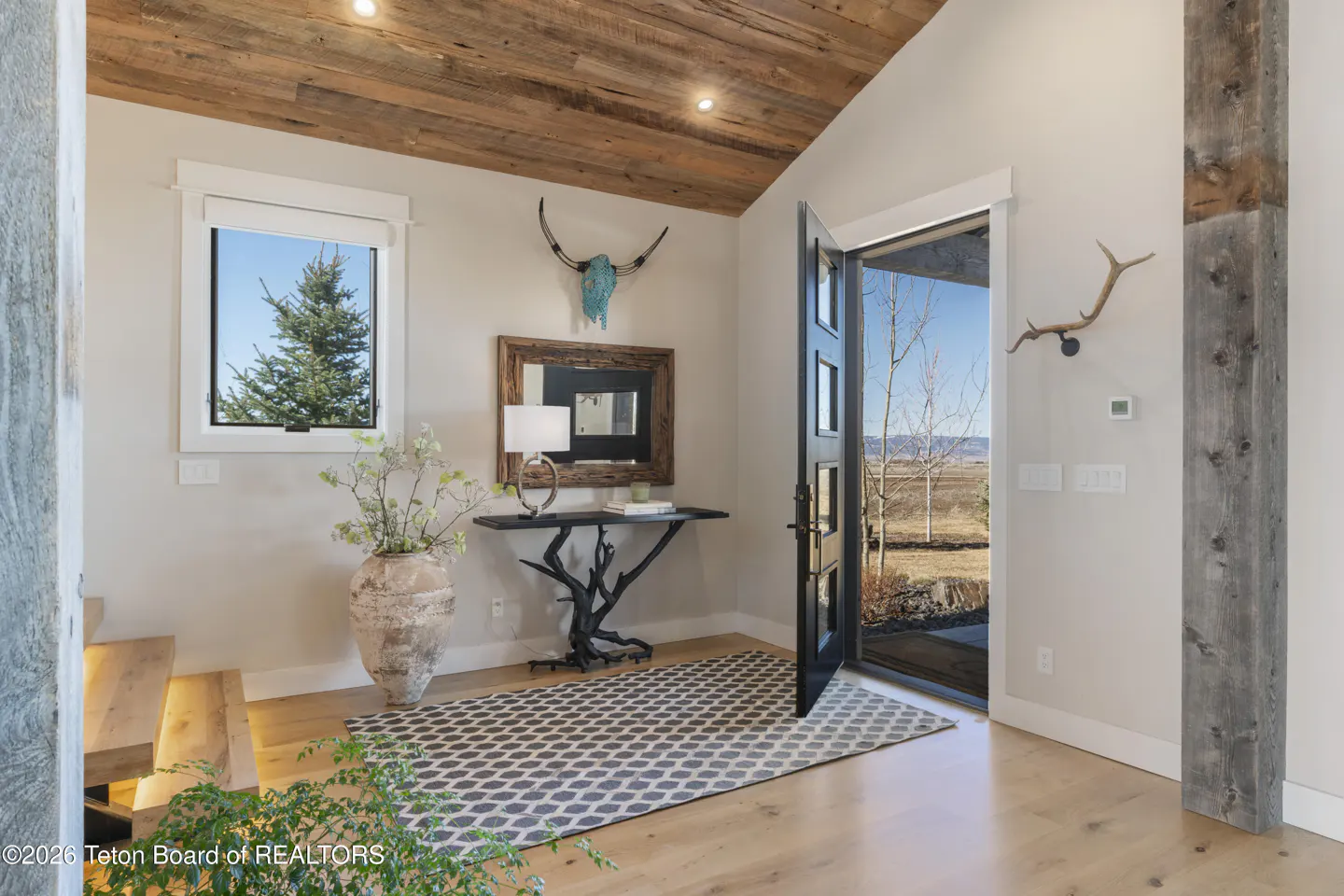 Entryway with wood ceiling, light walls, and wood floors. A black table with a lamp and mirror sits near a window. An open door reveals a landscape view.