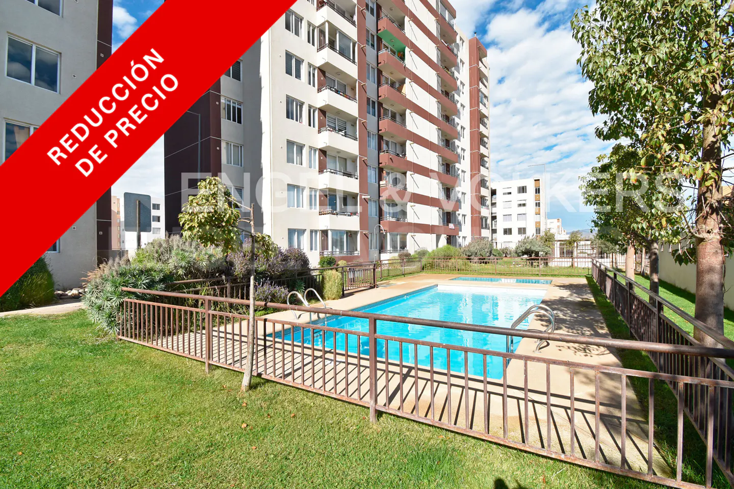 Apartment building with a pool. A red banner reads "REDUCCIÓN DE PRECIO". The pool is surrounded by a metal fence and grass.