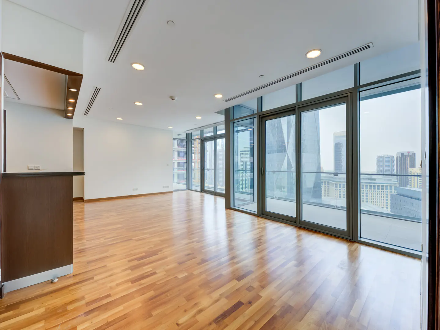 Bright, empty apartment with wood floors, white walls, and floor-to-ceiling windows showing a city skyline.