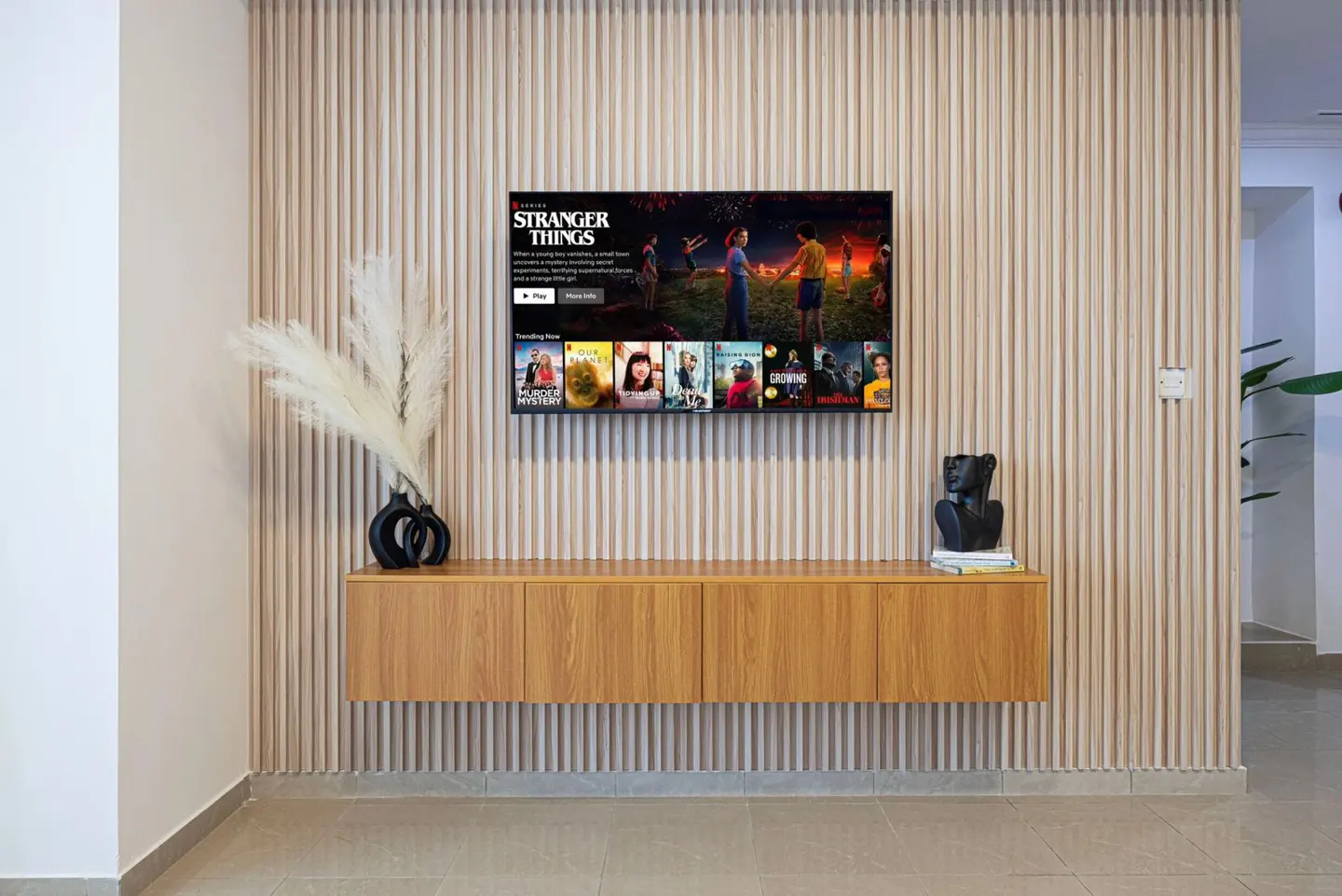Living room with a TV showing "Stranger Things" on a wood-paneled wall. A wooden cabinet sits below, decorated with a vase and sculpture.